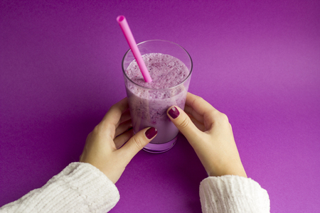 Female hands holds glass of berry smoothie with straw on purple background. Pink milkshake. Healthy drink. Blueberry cocktail.の写真素材