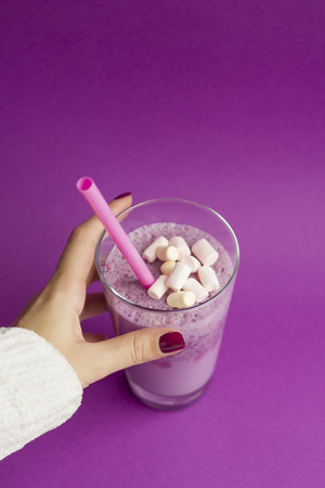 Female hands holds glass of berry smoothie with straw on purple background. Pink milkshake with copy space. Healthy drink. Blueberry cocktail and marshmallow.の写真素材