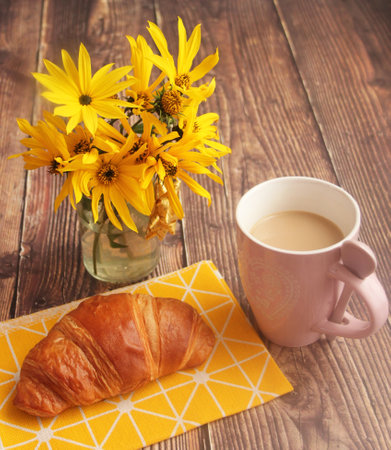 A cup of coffee with croissant and yellow flowers on a wooden backgroundの写真素材