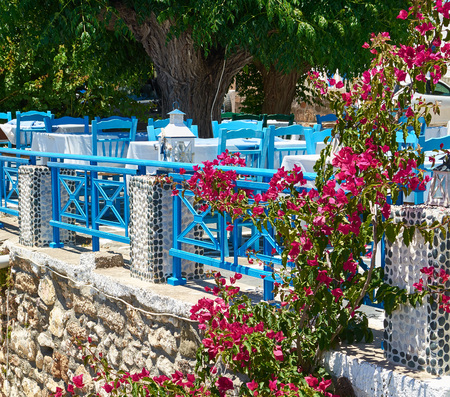 Terrace of summer restaurant in Greece, the island Rhodes. Blue chairs and tables with a white cloth under a big tree and the curling bush with bright purple flowers at a stone wall.の写真素材