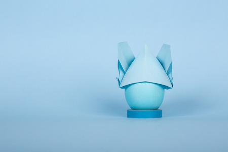 Happy Easter holiday concept dyed chicken egg in a bunny origami hat with ears on blue paper background with copy space.の写真素材
