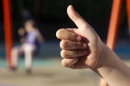 Child's hand showing thumb up and second kid on the background swings on the swings on the...
