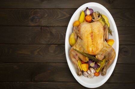 Whole roaster chikcken with backed vegetables in a dish on wooden dark brown background.