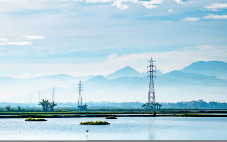Transmission tower, electricity pylon or a power pylon, standing majestically lined up to the horizon, in front of large lake or pond, also with beautiful cloud and mountain in the background, captured after sunriseの写真素材
