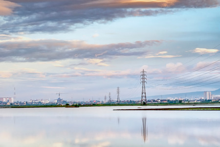 Transmission tower, electricity pylon or a power pylon, standing majestically lined up to the horizon, in front of large lake or pond, also with beautiful cloud and city in the background, captured at sunriseの写真素材