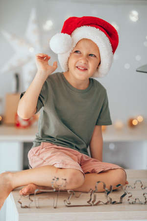 Boy in Santa hat is sitting on kitchen table and playing with cookie cutter.