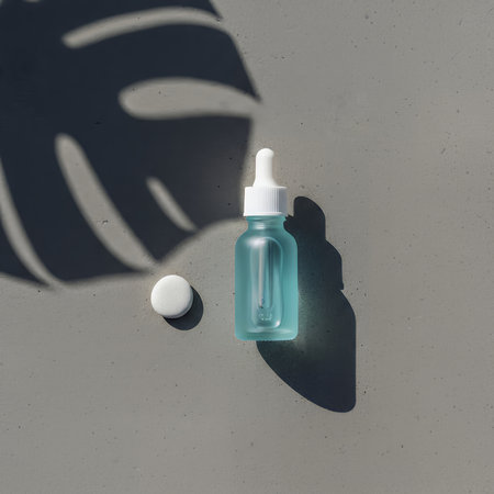 A blue frosted dropper bottle and a white pill are arranged on a grey background. Natural light casts a monstera leaf shadow, creating a minimalist and serene aesthetic.の素材