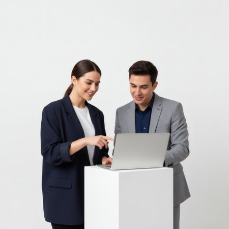 A businessman and businesswoman in professional attire work together on a laptop placed on a white pedestal against a clean studio background.の素材