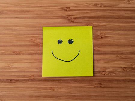 Sticker depicting the happy smiley character. Paper sticker pasted on a wooden surface. Self-adhesive paper sheet. A piece of paper on a wooden board. Reminder for action. Paper sheet for notes.の写真素材