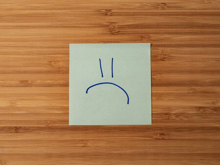 Sticker depicting the symbol of a sad emoticon. Paper sticker pasted on a wooden surface. Self-adhesive paper sheet. A piece of paper on a wooden board. Reminder for action. Paper sheet for notes.の写真素材
