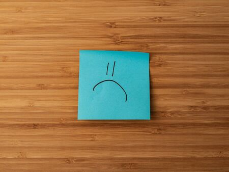 Sticker depicting the symbol of a sad emoticon. Paper sticker pasted on a wooden surface. Self-adhesive paper sheet. A piece of paper on a wooden board. Reminder for action. Paper sheet for notes.の写真素材