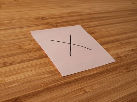 Sticker depicting the multiply symbol. Paper sticker pasted on a wooden surface. Self-adhesive paper sheet. A piece of paper on a wooden board. Reminder for action. Paper sheet for notes.の写真素材