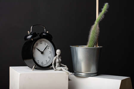 A woman sits leaning on an alarm clock. Clock on a plaster cube. Cactus in a metal pot.の写真素材