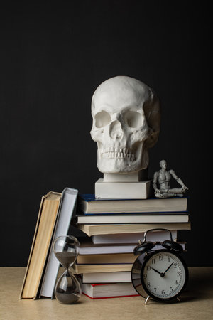 A man sits in a lotus position in the shadow of a plaster skull. Nearby are a clock showing 10 o'clock next to a stack of books. Composition in the dark key.の写真素材