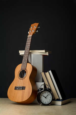 Ukulele next to books and clock showing 10 o'clock. Vertical composition in a dark key.