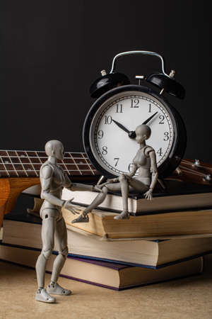 A woman sits on a stack of books next to an alarm clock. The ukulele neck is visible in the background. A man is talking to a woman, touching her knee. Composition in the dark key.の写真素材