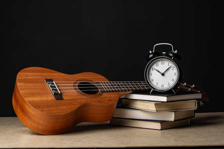 The ukulele lies on a stack of books. There is an alarm clock on a stack of books. Composition in the dark key.の写真素材