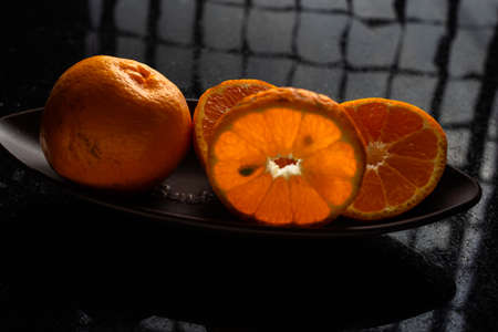 One whole tangerine and one cut in a brown canoe-shaped dish on a black stone table with a window reflection.の写真素材