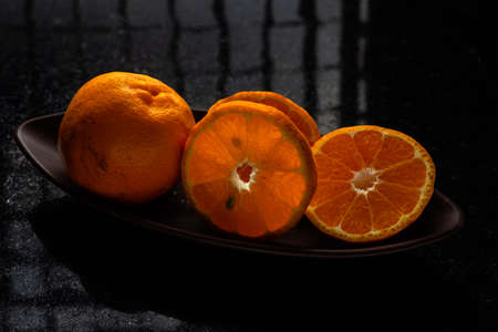One whole tangerine and one cut in a brown canoe-shaped dish on a black stone table with a window reflection.の写真素材