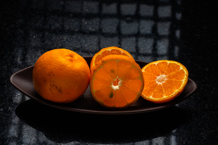 One whole tangerine and one cut in a brown canoe-shaped dish on a black stone table with a window reflection.の写真素材