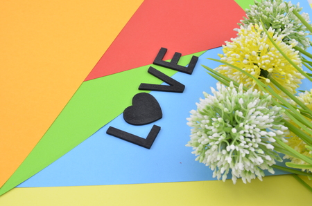word love with love symbol in black color.vertical position  with green, white and yellow artificial flower on orange, red, blue and green backgroundの写真素材