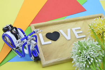 white word and black love symbol on colourfull background.green, yellow and white artificial flower placed on right. blue handcraft scooter on left sideの写真素材