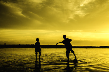image silhouette of two boys play beach soccer during sunset sunrise. blur on water and sky. reflect on waterの写真素材