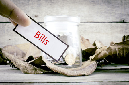 cropped image fingers holding white card with word BILLS. Coin in the glass jar surrounded with dry leaves with wood background.Financial concept with retro color themeの写真素材