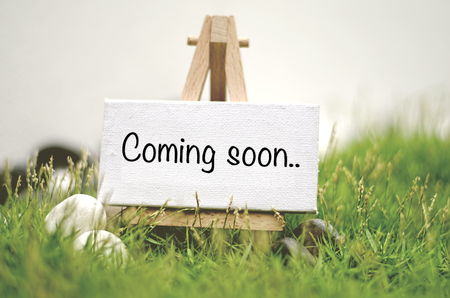 image concept white frame canvas on wooden tripod with word COMING SOON. Blurred and soft focus background with green grass and white stoneの写真素材