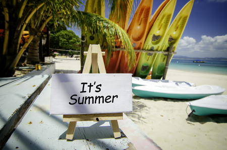 conceptual image with word IT'S SUMMER on white canvas frame and wooden easel. Blurred image of beach and colorful kayaks background at sunny day.の写真素材