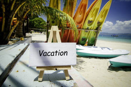 conceptual image with word VACATION on white canvas frame and wooden easel. Blurred image of beach and colorful kayaks background at sunny day.の写真素材