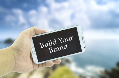 Conceptual image cropped hand holding mobile phone with word build your brand over blurred nature backgroundの写真素材
