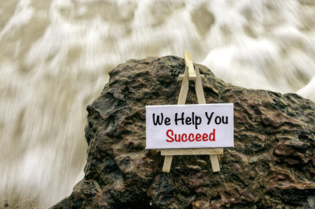 Image concept, word WE HELP YOU SUCCEED on white canvas frame with wooden tripod.background motion blur white wave flow hitting wet and mossy rock on the beach.の写真素材