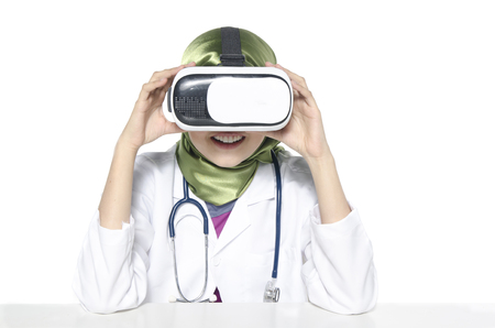Hijab woman doctor, veterinary or scientist with stethoscope sitting behind VR virtual reality goggles simulator isolated in white backgroundの写真素材