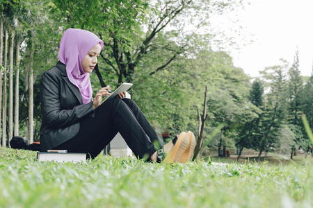 urban lifestyle,young business or muslimah student sitting on a green grass at park, reading an e-book on her tablet.の写真素材