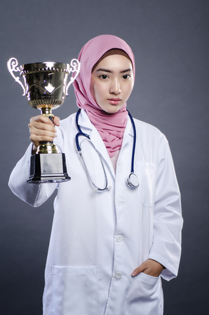 Symbol of success, closeup portrait of confident young hijab female doctor holding trophy over dark backgroundの写真素材