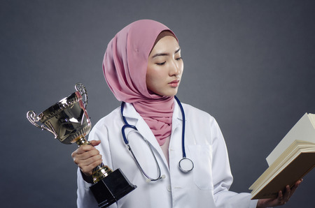 Symbol of success, closeup portrait of confident young hijab female doctor holding trophy and book over dark backgroundの写真素材