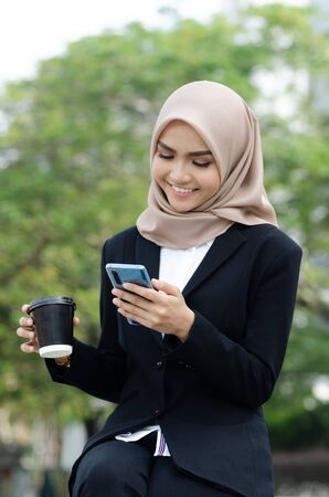 Portrait young businesswomen in black suit and hijab using smartphone and holding cup of coffeeの写真素材