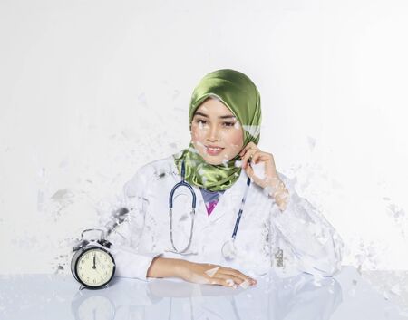 Young doctor with hijab. happy face expression siiting in front laptop ideal for stress management concept.vintage clock, green plant and book on table with reflectionの写真素材