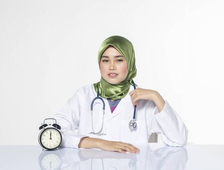 Young doctor with hijab. happy face expression siiting in front laptop ideal for stress management concept.vintage clock, green plant and book on table with reflectionの写真素材