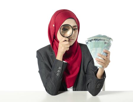 young woman in hijab looking on money through magnifying glass for authenticity checkingの写真素材