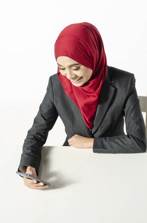 Communication concept, young woman with hijab sitting and texting her friends using her handphone against white backgroundの写真素材
