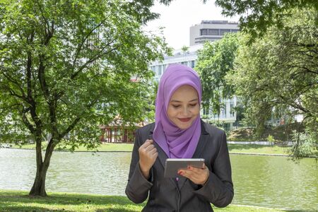 Communication concept, young woman with hijab sitting and texting her friends using her tablet at park during sunny dayの写真素材