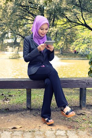 Communication concept, young woman with hijab sitting and texting her friends using her tablet at park during sunny dayの写真素材
