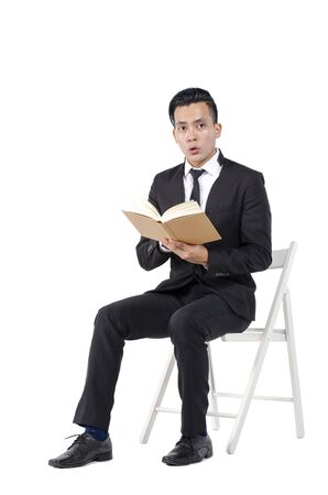 Portrait of young college student in black suit with books sitting against white backgroundの写真素材