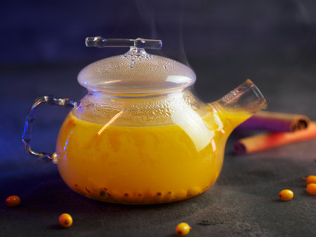 Tea with sea buckthorn in a glass teapot. Steam from hot tea. Colored backlight dark background. Seasonal hot berry warming drinkの写真素材