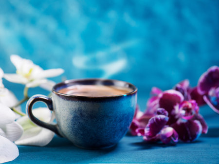 A blue cup of coffee on a blue table and a branch of a white and purple orchid.の写真素材