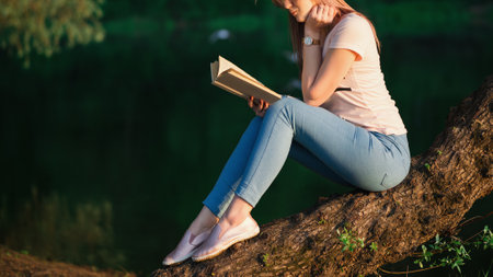 Young beautiful woman reading a book at sunset by the river. Read a book in silence in natureの写真素材