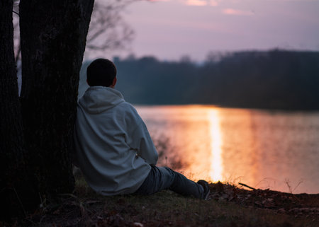 a lone young man in a white hoodie sits on the riverbank by a tree at sunsetの写真素材