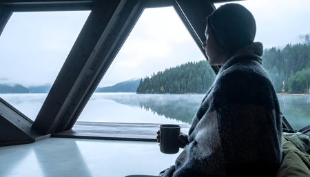 Man wrapped in a blanket, looking out the large A-frame window of a minimalist cabin at a foggy, mist-covered mountain lake landscapeの素材